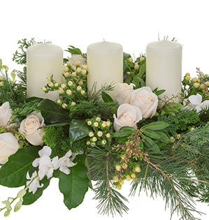 A holiday centerpiece in a ivory hues mixes Vendela roses, dendrobium orchids, pieris, Hinoki cypress, Deodar cedar, hypericum and fatsia leaves with white candles for a beautiful effect.