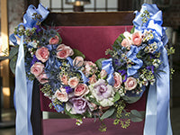 A floral chair swag is an elegant addition to a wedding, and this one is covered in a variety of pink and blush flowers and galax leaves, then it is finished with pale blue ribbons and bows on either end.