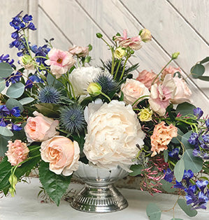 A compote design in the Pantone Color of the Year, Classic Blue, mixes blush Juliet garden roses, butterfly ranunculus, peonies, delphinium belladonna, lisianthus, eryngium, Italian ruscus, fatsia, pittosporum, and silver dollar eucalyptus.