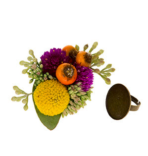 A small yet vibrant collection of orange rose hips, yellow crespedia, hot pink gomphrena, seeded eucalyptus, and sedum sits next to an antique brass ring blank.