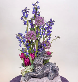 A square concrete base with a stone rabbit resting on one corner. Stems of Belladonna delphinium, veronica, allium, eryngium, and tulips are placed in a garden style behind the rabbit, with succulents tucked low next to the rabbits front paws.