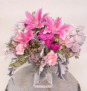 A large footed compote design with a base of dusty miller, waxflower and seeded eucalyptus, filled with carnations, spray roses, sweet peas, tulips, peonies, and oriental lilies.