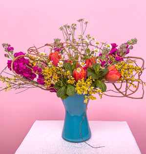 A vibrant blooming arrangement full of stock, genestra, tulips, feverfew and mint, paired with a vibrant aqua vase.