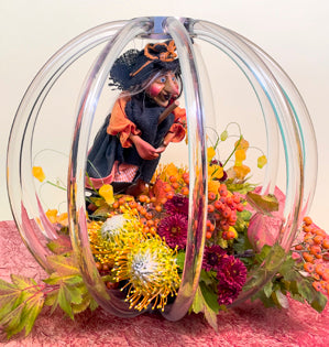 A Halloween floral design has a clear globe with a hanging witch and a base filled with vine maple, pin-cushion protea, mountain ash, spray mums, and sandersonia.