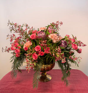 A mercury glass urn overflowing with a variety of evergreens, festival bush, spray roses, antique carnations, and protea.