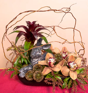 A lush and textural design made using a cement bunny, cymbidium orchids, pieris, scabiosa pods, bromeliad, sphagnum moss, and curly willow.