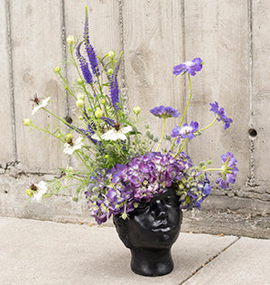 An ornate head vase is full of beautiful purple blooms like hydrangea, scabiosa, veronica, eryngium, and nigella.