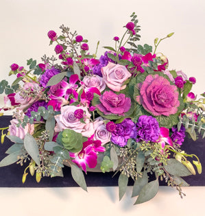 An abundant centerpiece in royal purple hues full of seeded eucalyptus, kale, gomphrena, Ocean Song roses, dendrobium orchids, and carnations.