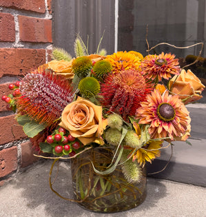 A glass vessel with curly willow wrapping around the inside to create a rooted effect, and then filled with Banksia, toffee roses, coneflower blooms and pods, hypericum berries, and millet all in muted fall tones.