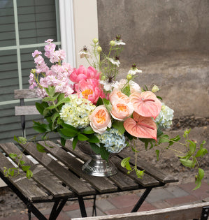 A blooming round arrangement in the soft Bespoke style uses hydrangeas, peonies, anthurium, garden roses, and stock mixed with a bit of nigella, ruscus, mint and clematis vine draping down.