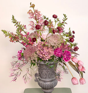 A large urn overflowing with, Italian ruscus, hydrangea, miniature cymbidium orchids, tulips, foxglove, Queen Anne's lave, carnations, Westminster roses, jasmine vine, and butterfly ranunculus.