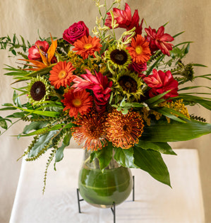 A large lush vase arrangement in bold red and orange tones features pin cushion protea, roses, Gerbera daisies, sunflowers, Bird of Paradise, banksia, solidaster, kangaroo paws and palm fronds.
