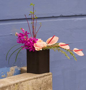 A beautiful linear design combines anthurium, pink Mondial roses, Aranda orchids, lily grass, red twig dogwood, and knife acacia in a modern pottery container.