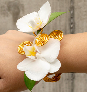 A close-up of a wrist wearing a gold wire cuff bracelet decorated with white orchid flowers and green leaves, shown against a blurred gray background.