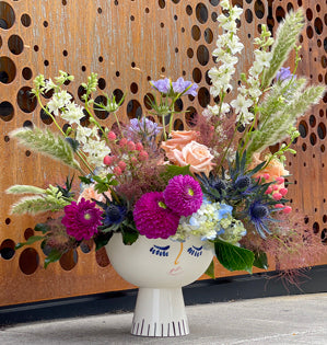 A colorful floral arrangement in a white ceramic pedestal vase, set against a brown wooden slat wall with circular cutouts.