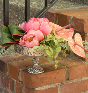 A decorative floral arrangement featuring pink peonies, pink and red anthuriums, and green foliage in a silver pedestal bowl, displayed on a brick ledge.