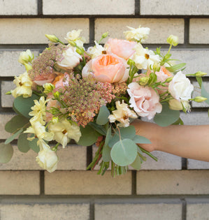 A soft pastel floral bouquet held by a person's hand against a light beige brick wall. The bouquet features peach roses, cream lisianthus, dusty pink filler flowers, and green eucalyptus leaves.