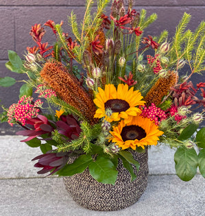 A vibrant autumnal floral arrangement in a textured gray woven basket, featuring bright yellow sunflowers, red berries, brown grasses, and green foliage against a dark gray brick wall.