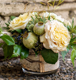 A rustic floral arrangement featuring cream-colored roses, green poppy pods, and green leaves in a small glass jar vase, set on a gravel surface.