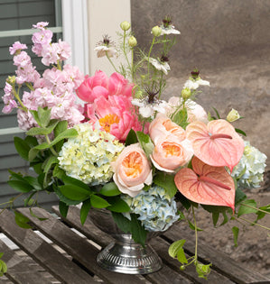 A vibrant artificial flower arrangement in a silver pedestal vase, featuring pink peonies, white daisies, pink and white anthurums, and light blue hydrangeas on a weathered wooden table.