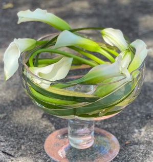 White calla lilies with green stems arranged inside a clear glass pedestal dish on a gray stone surface.