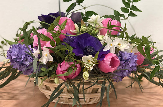 A spring floral arrangement in purple, white and pink color palette features spring flowers such as hyacinth, anemones, paperwhites, tulips and jasmine vine.