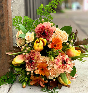 A beautiful wedding bouquet in a peach, orange and cream color palette features spray roses, tulips and ranunculus accented with lacquered bay leaves and maiden hair fern.