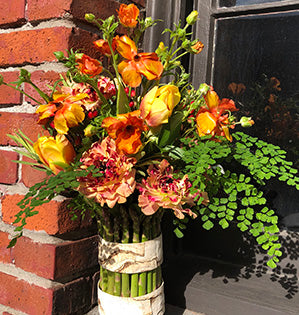 A fresh springtime vase arrangement in orange and yellow colors combines fresh asparagus, Japanese ranunculus, butterfly ranunculus, tulips, hypericum, and a variety of foliages.