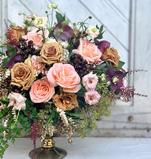 A compote centerpiece features Toffee roses, Oceania roses, peach carnations, hypericum, astilbe, butterfly ranunculus, hellebores, and andromeda.