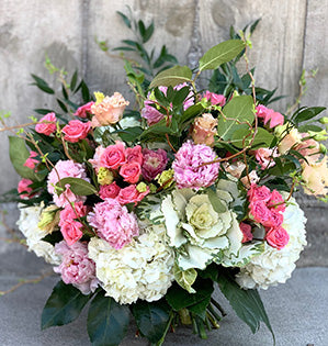 A hand-tied garden style bouquet features white hydrangea, kale, pink peonies, spray roses, and peach colored lisianthus for a beautiful effect.