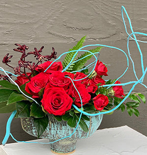A contemporary floral design in red and turquoise hues mixes painted curly willow with standard roses and spray roses, kangaroo paws, and a variety of foliage, all in a beautiful turquoise compote.