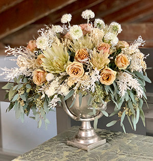A fabulous mix of fresh, dried, and preserved flowers and foliage such as protea, roses, scabiosa, ruscus, and eucalyptus in cream, mustard and toffee colors is set into a large silver compote for a beautiful effect.