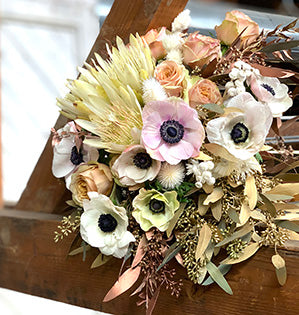 A crescent style hand-tied bouquet features white protea, roses, anemones, dried and bleached flowers, and is enhanced with a collar of metallic copper and gold foliages.