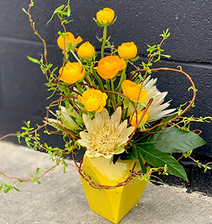 A vibrant floral design features beautiful yellow ranunculus, protea, fatsia, and an armature of curly willow.