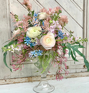An arrangement for the home features blue tweedia, fragrant stock, marvelous white ranunculus, sweet peas, dried and bleached ruscus, blue star fern, and jasmine vine.