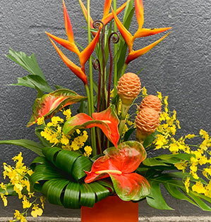 A beautiful tropical floral arrangement in orange and yellow colors features a collection of heliconia, oncidium orchids, anthurium, fern curls, beehive ginger, and monstera leaves.