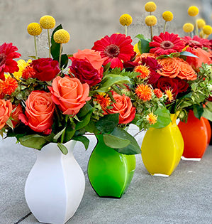 Flower arrangements in four different colored vases: white, green, yellow and orange combine spray roses, Gerbera daisies, craspedia,  safflower, Israeli ruscus, leather fern, and salal. 
