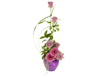 A vertical line design arrangement of soft lavender roses and light pink Gerberas accentuated with two curved strands of long grass, ruscus and galax foliage, and with Spanish moss hanging over the edges of a metallic purple container.
