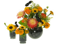 A trio of arrangements, one in a large bubble bowl and two in smaller hurricane vessels, featuring dahlias, marigolds, mini sunflowers, roses, and solidaster in whimsical oranges and yellows with little black bats that appear to be hovering above.