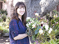 Amy Osaba stands holding a bouquet of foraged flowers and foliage. Amy is wearing a blue dress with thin red windowpane plaid detail.