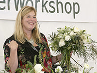 Holly Chapple smiles while holding her own creation of a loose, airy, wispy bouquet of white roses with other cascading ingredients like plumosa, seeded eucalyptus, white lilac, and feather willow yew.