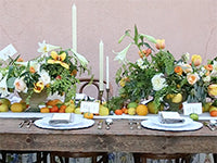 A dining table has two place settings, three long candlesticks, two large floral arrangements of lilies, tulips, garden roses, and an abundance of accompanying foliage, and all along the table runner sit a variety of citrus.