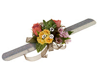 A corsage containing coral garden roses, pink wax flowers, yellow spray roses, and a small succulent with a chocolate-colored ribbon and three diamante pin accents and is displayed.