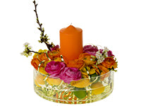 A round dish holds a bed of colorful ranunculus in pink, orange, and yellow surrounding an orange candle. Lemons and limes can be seen submerged bellow. White wax flowers and curled gold flat wire adorn the arrangement. 