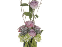 A closeup of a tall, striking arrangement showing green hydrangea, soft lavender roses, silver gilded succulents, sparkling branches, and hanging amaranthus.