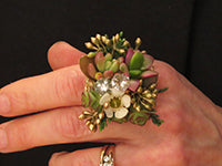 A hand against a black background wears two rings: one a gold and diamond wedding band and the other created from small succulents, white wax flower, gilded seeded eucalyptus, evergreen, and white diamante gems.