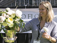 Kim Curtis smiles as she works on an arrangement of garden roses and foliage. Kim is wearing a gray shirt and a name card lanyard around her neck.