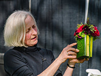 Francoise Weeks looks at a compact arrangement of blooms in bold fuschia and purple and various foliages she is holding. Francoise is in a black shirt and is pointing at a detail of the arrangement.