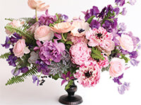 A fun arrangement of peonies, ranunculus, and other blooms in soft pinks and purples complimented with sword ferns, succulents, and dusty miller. The skinny foot of the container can be seen below the full arrangement.
