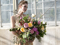 A woman with brown hair pulled up in an updo and wearing a nude-colored dress looks away somberly while holding a large and impressive bouquet of peonies in various colors nestled with severals kinds of foliage.
